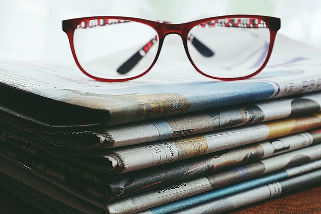 Auf dem Foto abgebildet ist eine rote Brille, die auf einem Stapel Zeitungen liegt. 