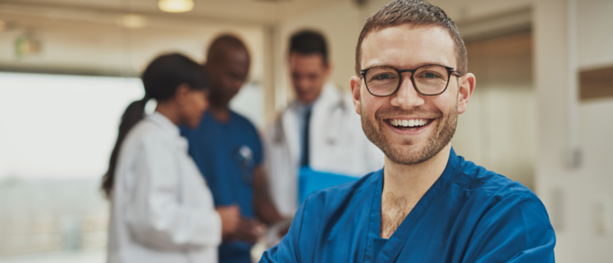Abgebildet ist ein junger Mann mit Brille in seiner Arbeitskleidung als Pflegefachmann, der in die Kamera lacht.