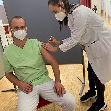 Eine Ärztin impft einen Pfleger. Beide tragen FFP2-Masken.