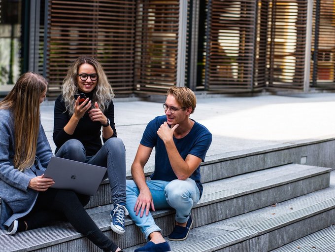 Abgebildet sind drei Personen, die mit Handy und Laptop auf Treppenstufen sitzen und sich austauschen.