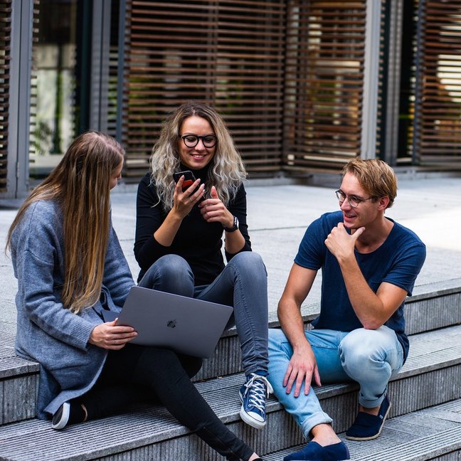 Abgebildet sind drei Personen, die mit Handy und Laptop auf Treppenstufen sitzen und sich austauschen.