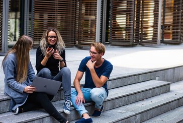 Abgebildet sind drei Personen, die mit Handy und Laptop auf Treppenstufen sitzen und sich austauschen.